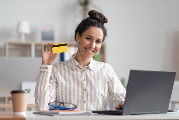 Portrait de femme d'affaires heureuse tenant la carte de crédit tout en faisant des achats en ligne sur ordinateur portable et souriant à la caméra - Photo, image