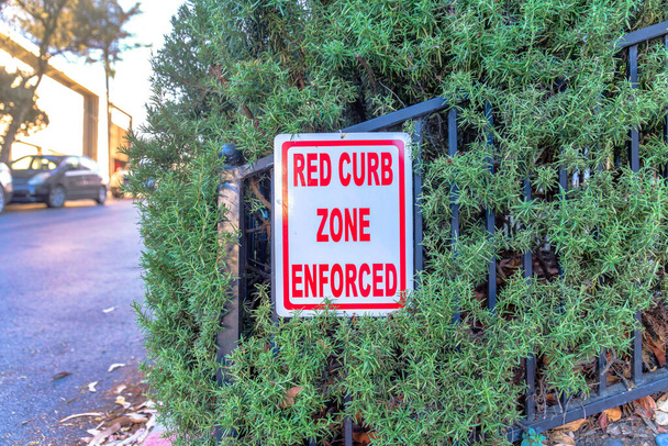 Czerwony krawężnik wymuszony znak przy drodze w San Francisco w Kalifornii. Oznakowanie umieszczone na metalowym ogrodzeniu, przyklejone do zielonych krzewów i zaparkowanych pojazdów na drodze z tyłu. - Zdjęcie, obraz