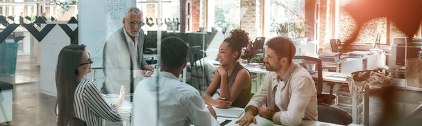 Spotkanie biznesowe. Dojrzały biznesmen omawia coś ze swoimi młodymi kolegami, siedząc razem przy stole biurowym w nowoczesnym biurze - Zdjęcie, obraz