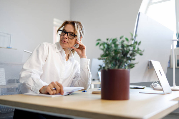 Secretária adulta bonita em uma camisa branca e óculos senta-se no local de trabalho - Foto, Imagem