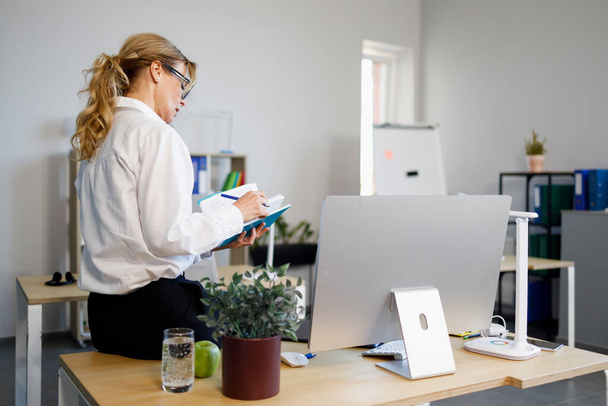 Элегантная деловая женщина сидит в офисе за столом с блокнотом и ручкой в руках - Фото, изображение