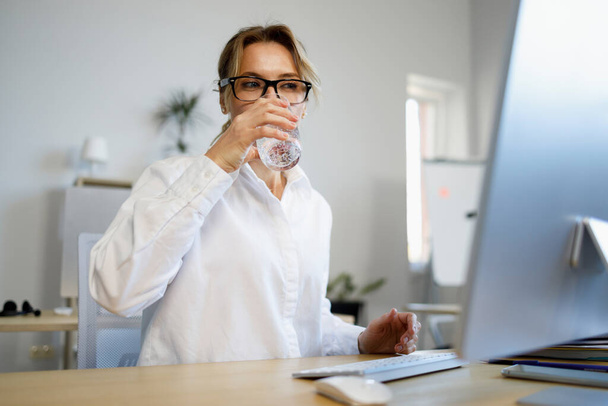 Όμορφη ώριμη επιχειρηματίας που εργάζεται σε έναν υπολογιστή και πόσιμο ποτήρι γλυκό νερό  - Φωτογραφία, εικόνα