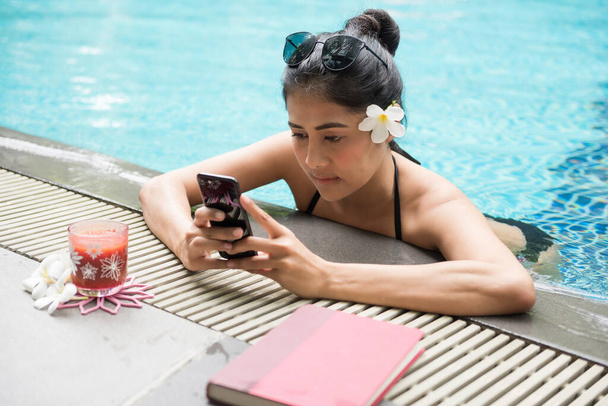 Belle femme asiatique en bikini noir jouer smartphone par bord de piscine. Vacances d'été. Chat, SMS, médias sociaux en ligne, achats en ligne par téléphone mobile - Photo, image