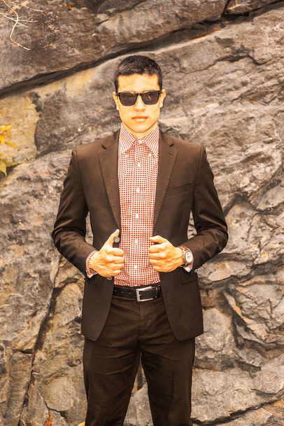 Attitude. Dressing in black suit, red, white patterned undershirt, wearing sunglasses, wristwatch, short hair cut, a young sexy guy standing by rocky wall, mysteriously looking at yo - Photo, Image