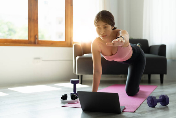 gesunde asiatische Sportlerin in Sportbekleidung zu Hause mit Laptop, um Trainingsergebnisse in den sozialen Medien zu teilen und Bauchmuskelübungen auf Fitnessmatte zu machen - Foto, Bild
