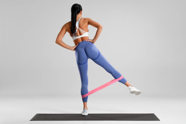 Vücut geliştiren kadın, gri arka planda direnç bandıyla kalça egzersizi yapıyor. Atletik kız spor yapıyor. - Fotoğraf, Görsel