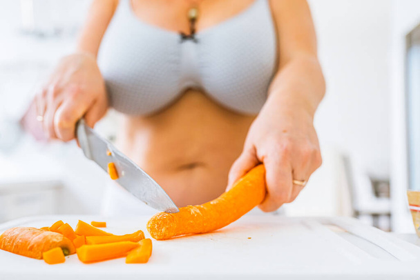unrecognizable woman in lingerie, bikini and bra, with curvaceous figure, cuts fresh carrots with large chef's knife. Attractive woman in bikini cooking vegetables in kitchen - Φωτογραφία, εικόνα