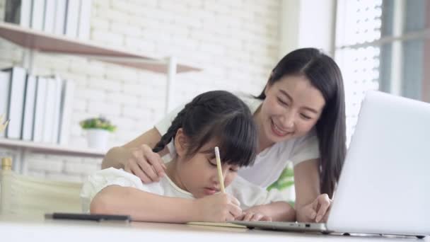 Nauka w domu. Szczęśliwa młoda Azjatka matka i córka odrabiają lekcje przy laptopie z pomocą matki, siedząc na biurku w domu. Dziewczyna z radością uczy się w domu. Koncepcja edukacji internetowej - Materiał filmowy, wideo