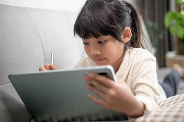 Dziewczynka korzystająca z tabletu w Internecie, dziecko siedzące na kanapie oglądające lub rozmawiające z przyjacielem online, dziecko relaksujące się rano w salonie, dzieci z koncepcją nowej technologii - Zdjęcie, obraz