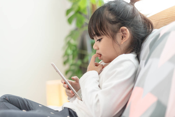 Cute little kid girl watching video on smartphone with smiley face alone on the bed, child using mobile phone with happy face at home. Stay at home quarantine coronavirus COVID-19 pandemic prevention. - 写真・画像