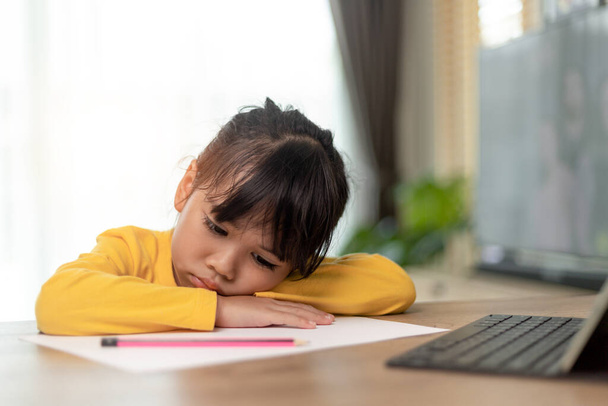 小さなアジアの女の子一人で座って退屈そうな顔で外を見ると、就学前の子供は宿題で悲しい退屈とテーブルの上に頭を置く、甘やかされた子供 - 写真・画像