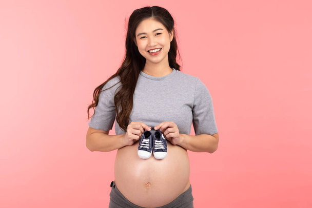 Azjatka w ciąży trzyma trampki dla noworodka odizolowane na różowym tle.Kobieta w ciąży pakowania rzeczy dla dzieci gotowy do szpitala położniczego.Ciąża Przygotuj się na noworodka Concept - Zdjęcie, obraz