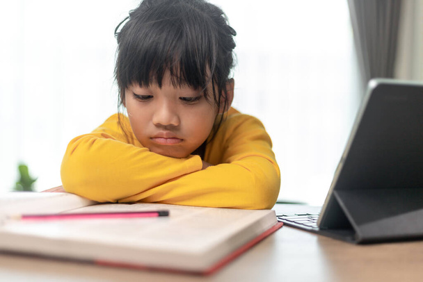 Kis ázsiai lány ül egyedül, és néz ki egy unatkozó arc, óvodás gyermek feküdt fejét az asztalra szomorú unatkozik a házi feladatot, elkényeztetett gyermek - Fotó, kép