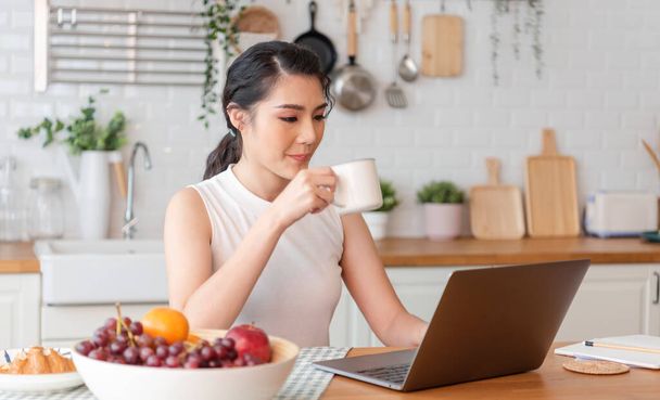 Beautiful young asian woman working on laptop computer while sitting at the kitchen room, drinking coffee. - Photo, image