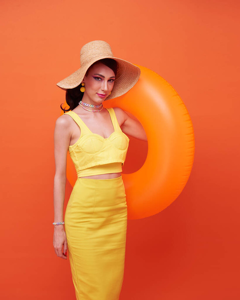 молода усміхнена весела туристична жінка в капелюсі тримає надувний кільце ізольовано на помаранчевому фоні
. - Фото, зображення