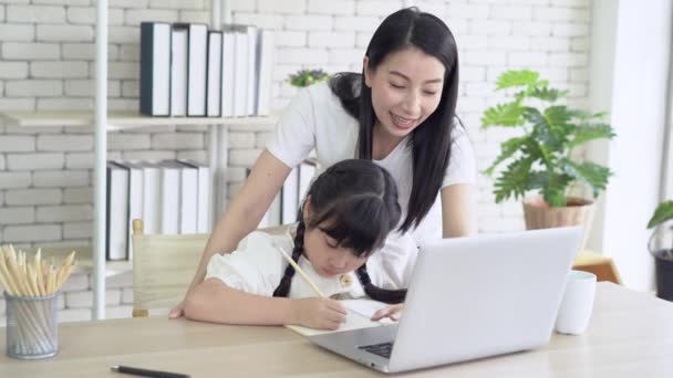 Porträt einer jungen asiatischen Mutter, die ihrer Tochter bei den Hausaufgaben hilft, während sie zu Hause am Schreibtisch sitzt. Mutter flechtet Tochter Haare und lächelt. Kleines Mädchen freut sich, zu Hause zu lernen. - Filmmaterial, Video