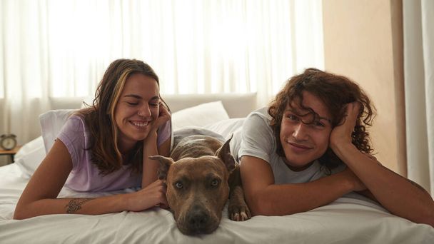 Young joyful caucasian couple rest with dog on bed - Foto, Bild