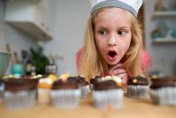 My first batch of cupcakes. Shot of a surprised little girl looking at cupcakes she baked at home. - Valokuva, kuva