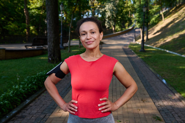 Bájos izmos nő egészséges esztétikai testtel és ragyogó bőrrel, sportoló piros pólóban és okostelefon tartó pózol karokkal derékon nézi a városi park futópadján álló kamerát. - Fotó, kép