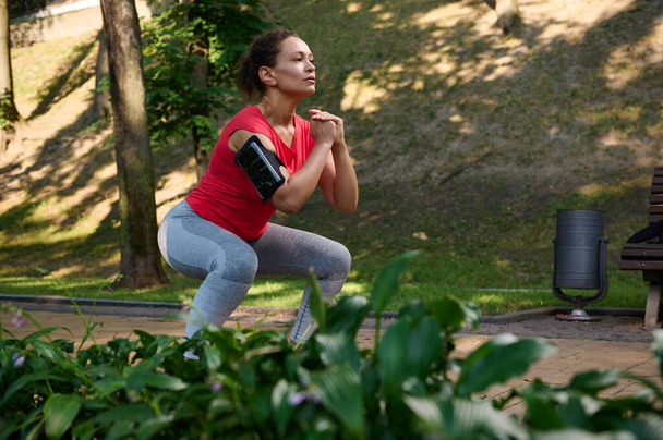 Determined athlete, sportswoman exercising outdoor, doing deep squats in the city park. Enjoy summer days and keep your body fit. Sport, fitness, functional body weight training concept - 写真・画像