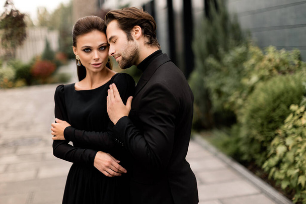 Portrait of fashion young adult couple on the photoshoot. Handsome beard man in stylish suit hugging gorgeous woman in black dress, posing for the photo. - 写真・画像