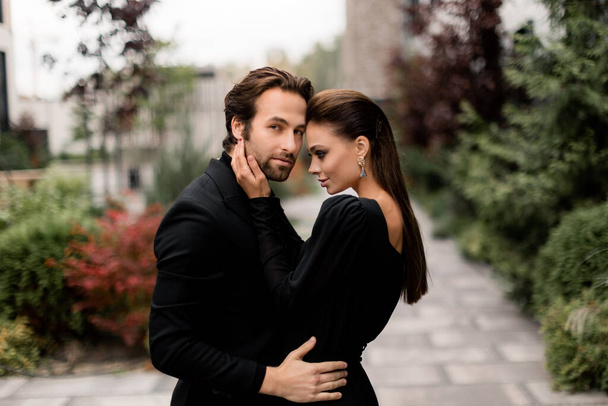 Portrait of fashion young adult couple on the photoshoot. Handsome beard man in stylish suit hugging gorgeous woman in black dress, posing for the photo. - Foto, imagen