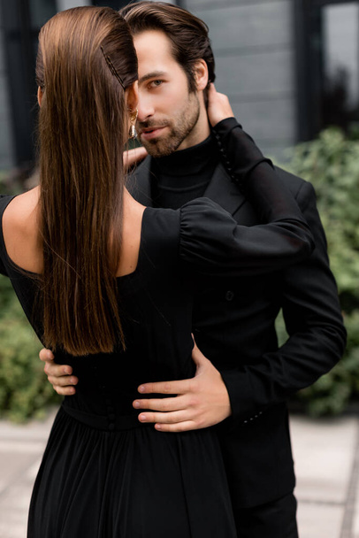 Portrait of fashion young adult couple on the photoshoot. Handsome beard man in stylish suit hugging gorgeous woman in black dress, posing for the photo. - Fotografie, Obrázek