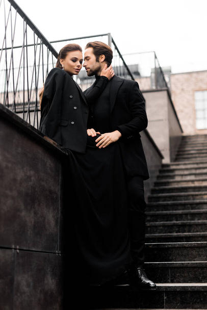 Portrait of fashion young adult couple on the photoshoot. Handsome beard man in stylish suit and gorgeous woman in black dress posing on the urban stairs. - Valokuva, kuva