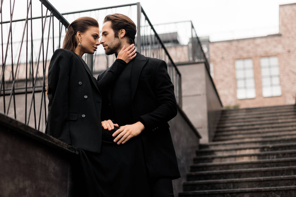 Portrait of fashion young adult couple on the photoshoot. Handsome beard man in stylish suit and gorgeous woman in black dress posing on the urban stairs. - Фото, изображение