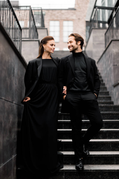 Πορτρέτο της μόδας νεαρό ζευγάρι ενηλίκων στη φωτογράφηση. Όμορφος γενειοφόρος με κομψό κοστούμι και πανέμορφη γυναίκα με μαύρο φόρεμα που ποζάρει στις σκάλες της πόλης. - Φωτογραφία, εικόνα