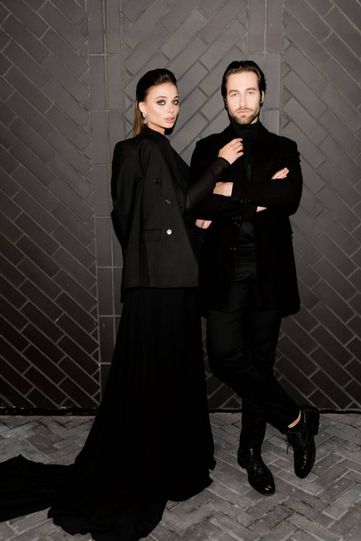 Stylish dressed caucasian couple having a photoshoot for magazine cover in black outfits. Beautiful young woman with charming beard man posing at the camera, fashion models concept.  - Foto, Imagem