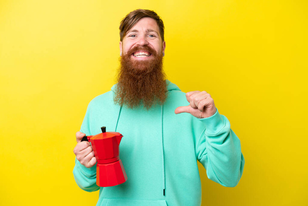 Redhead man with beard holding coffee pot isolated on yellow background proud and self-satisfied - 写真・画像
