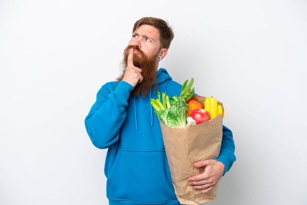 Rothaariger Mann mit Bart, der eine Einkaufstasche auf weißem Hintergrund hält und beim Nachschauen Zweifel hat - Foto, Bild