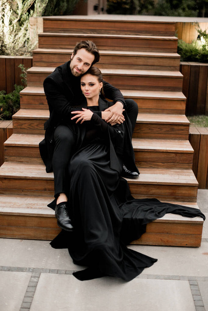 Portrait of fashion young adult couple on the photoshoot. Handsome beard man in stylish suit and gorgeous woman in black dress sitting on wooden stairs.  - 写真・画像