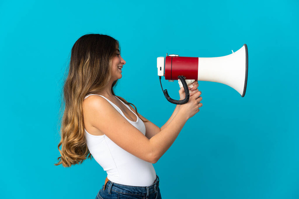 Young caucasian woman isolated on blue background shouting through a megaphone to announce something - Foto, afbeelding