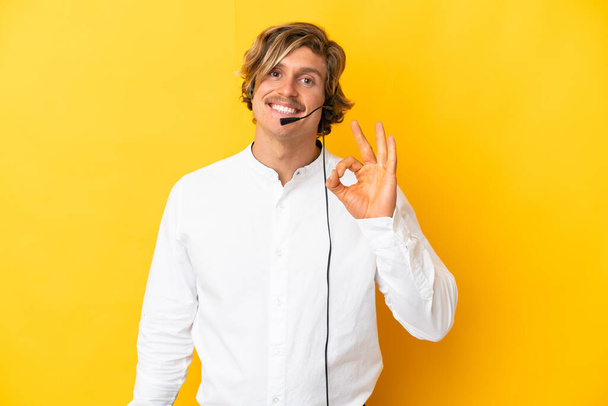 Telemarketer man working with a headset isolated on yellow background showing ok sign with fingers - Фото, изображение