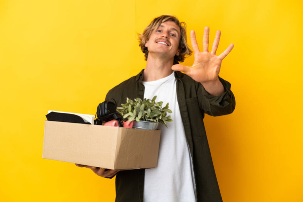 hombre rubio haciendo un movimiento mientras recoge una caja llena de cosas aisladas sobre fondo amarillo contando cinco con los dedos - Foto, imagen