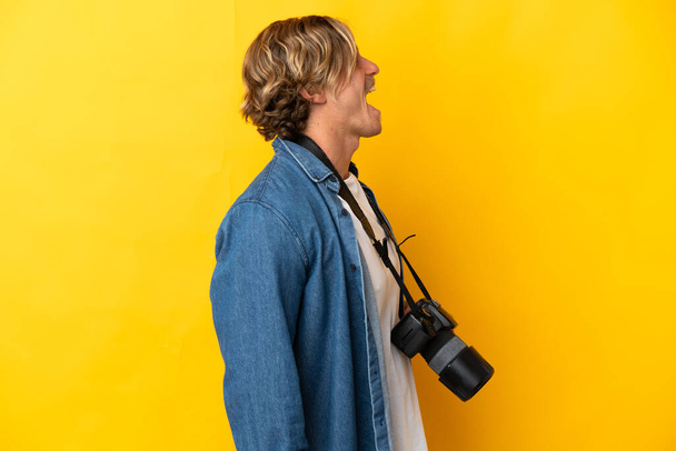 Jonge fotograaf man geïsoleerd op gele achtergrond lachen in laterale positie - Foto, afbeelding