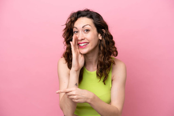 Young caucasian woman isolated on pink background pointing to the side to present a product and whispering something - Фото, зображення