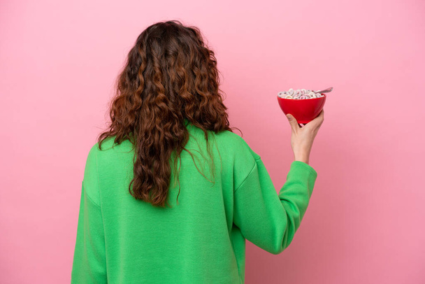 Young caucasian woman holding a bowl of cereals isolated on pink background in back position - Foto, immagini