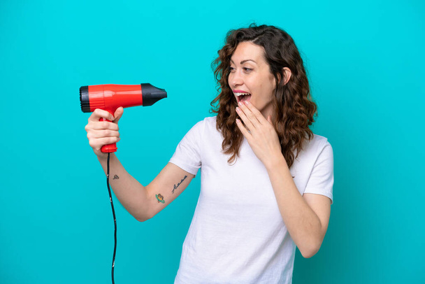 Young caucasian woman holding a hairdryer isolated on blue background with surprise and shocked facial expression - Foto, imagen
