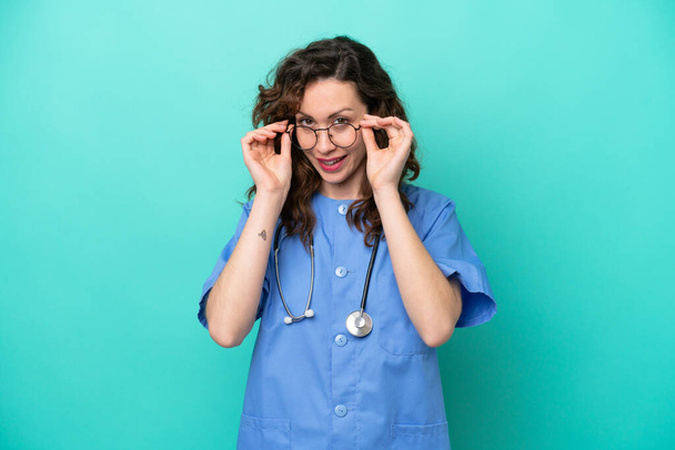 Νεαρή νοσοκόμα καυκάσια γυναίκα απομονωμένη σε μπλε φόντο με γυαλιά και έκπληκτος - Φωτογραφία, εικόνα