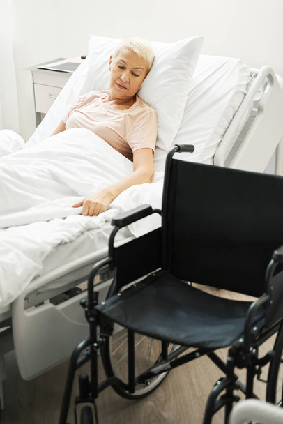 Ηλικιωμένος πολίτης στο κρεβάτι κοιτάζοντας αναπηρική καρέκλα - Φωτογραφία, εικόνα