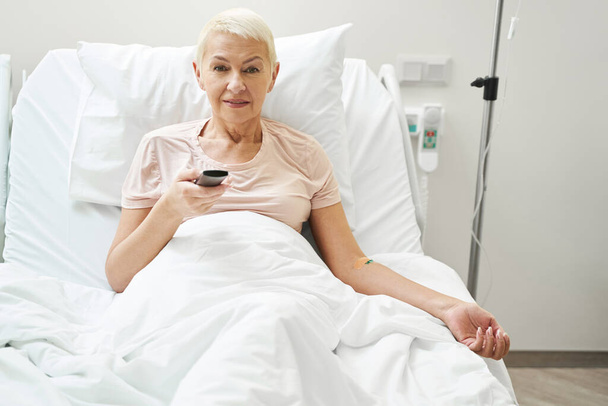 Paciente de edad avanzada que hace clic en TV remota mientras está bajo terapia intravenosa - Foto, Imagen