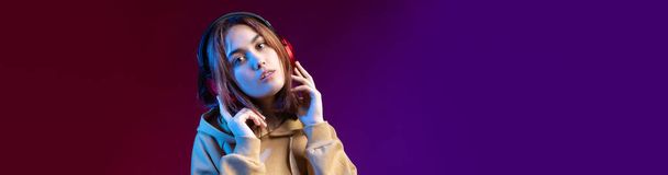 Joven chica hipster hermosa de moda vestida con una sudadera con capucha escuchando música en auriculares rojos en un estudio fondo dramático rojo - Foto, imagen