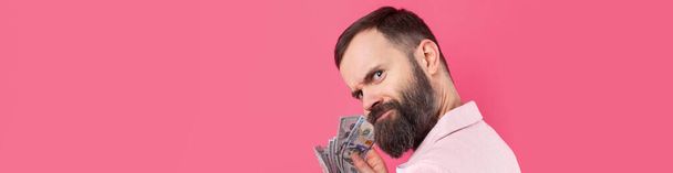 ピンクのジャケットを着た髭を生やした若いビジネスマンの肖像画は、赤いスタジオの背景に$紙幣を示しています。味、お金の匂い. - 写真・画像