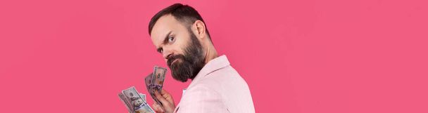 Portrait of a contented young businessman with a beard dressed in a pink jacket showing us dollar banknotes against a red studio background. Taste, smell of money. - Foto, Imagem