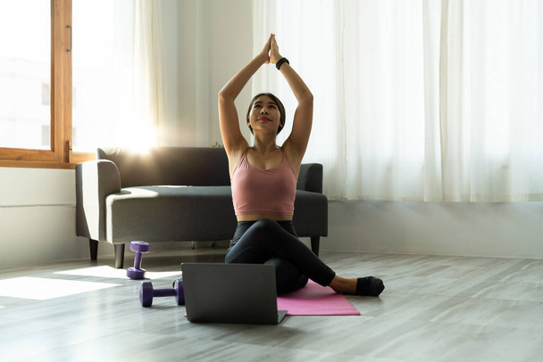 Calm relaxed young asian woman sitting in lotus position with hands up namaste gesture, closed eyes, breathing, resting, deeply meditating, practicing yoga on yoga mat at home - Foto, imagen