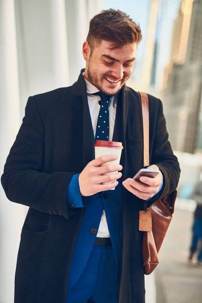 Starting the day right. Shot of a cheerful young man texting on his phone while drinking coffee in the busy streets of the city while going to work in the morning. - Foto, Imagen
