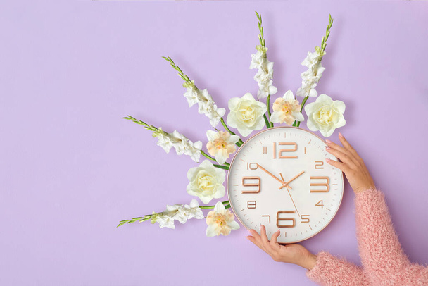 Female hands with clock and blooming flowers on lilac background. Daylight saving time - Photo, Image